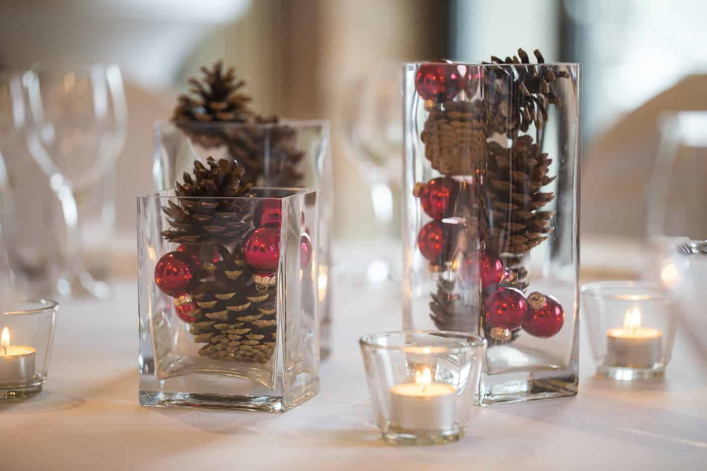 Zwei Glasvasen mit Tannenzapfen und roten Beeren neben Teelichtern