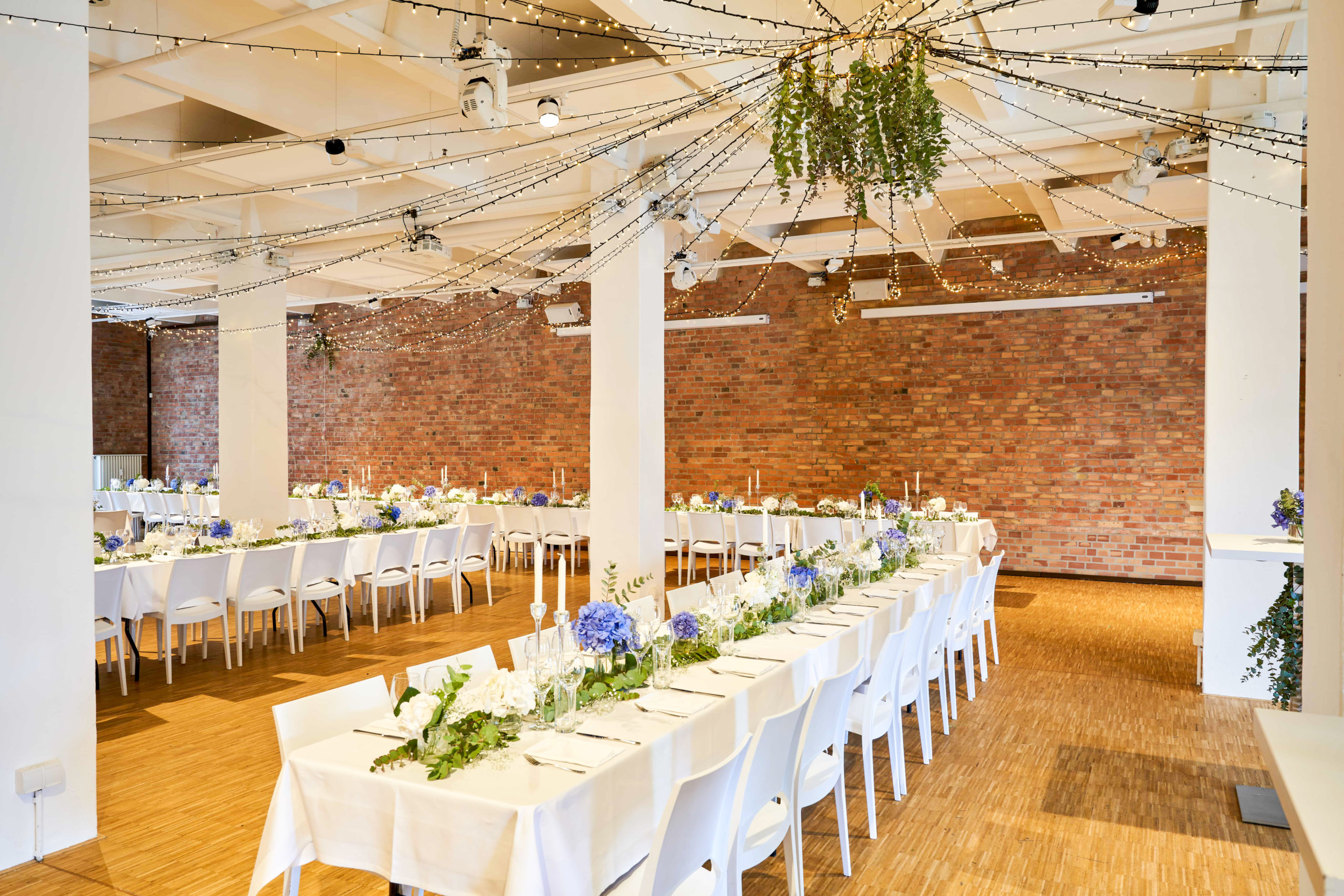 Festlich gedeckte lange Hochzeitstafel im Backsteinloft mit weißen Stühlen und Lichterketten